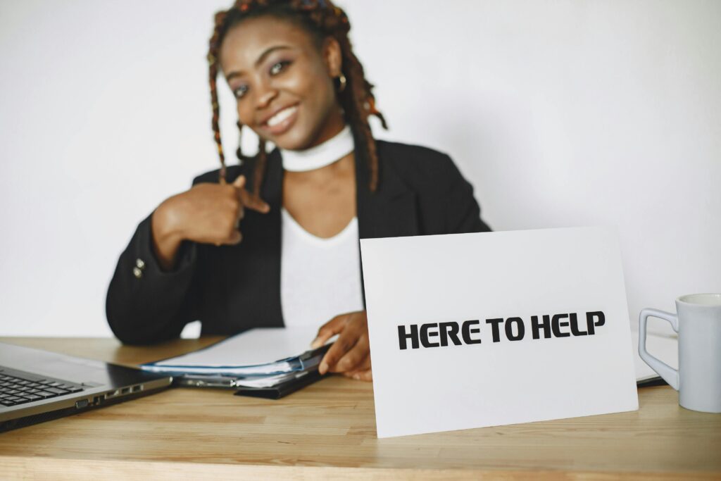Smiling businesswoman ready to assist, sitting at desk with helpful sign.